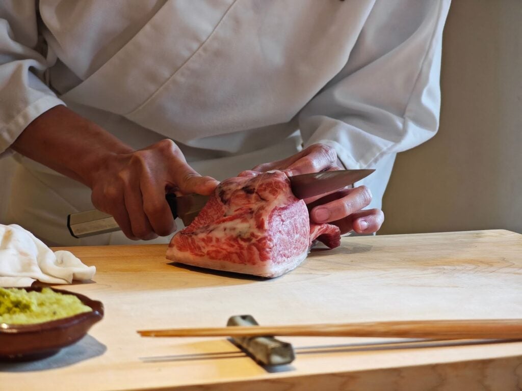 Chef corta a cuchillo una pieza de wagyu sobre tabla de madera, con wasabi y palillos, en una experiencia de estilo omakase.