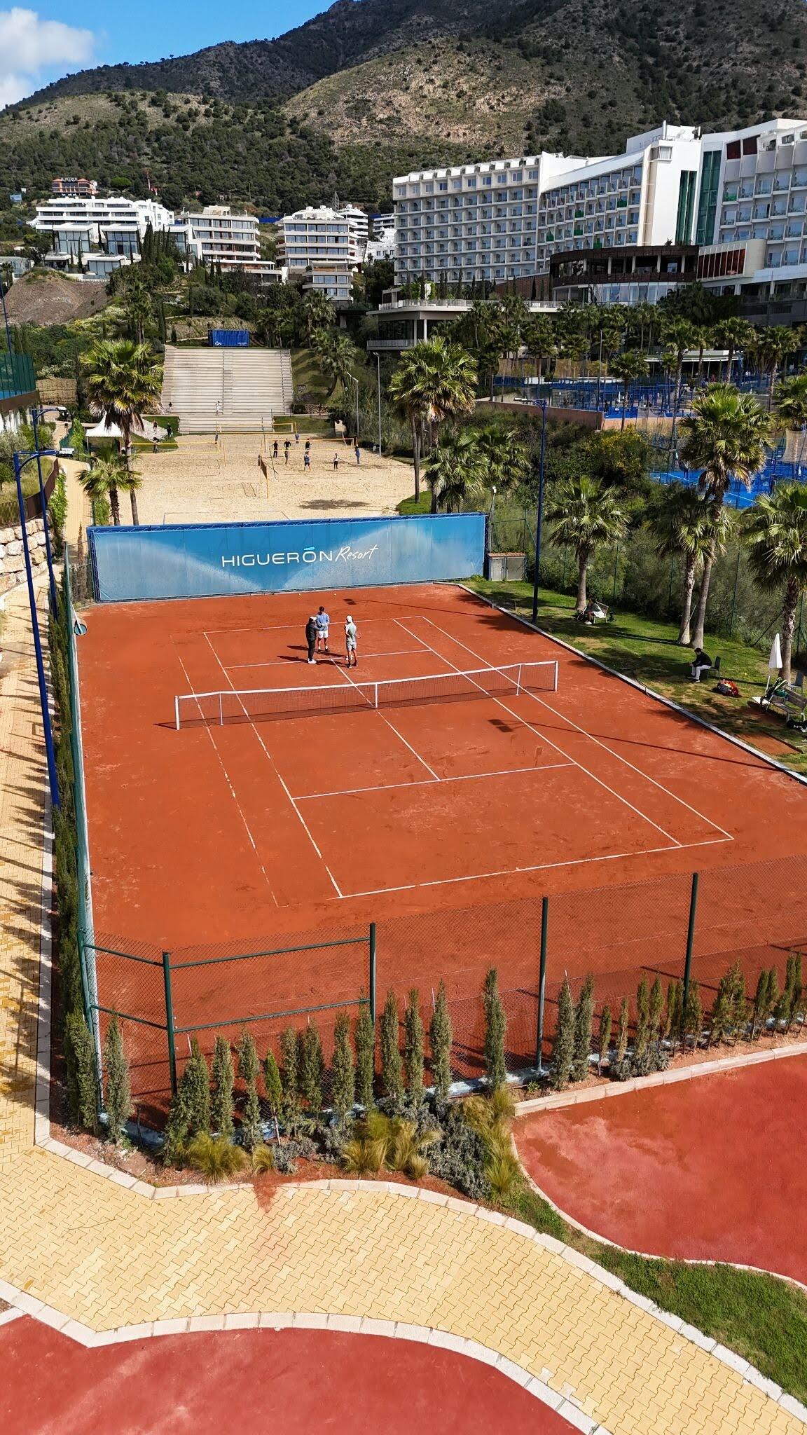 Pista de tenis de tierra batida del Higuerón Tennis Center, con jugadores y el resort al fondo, en la Costa del Sol