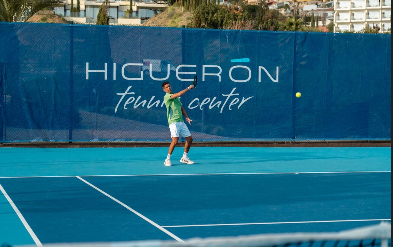 Jugador de tenis golpeando la pelota en una pista azul de Higuerón Tennis Center.